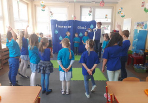 Dzieci ubrane na niebiesko stoją w kole z nauczycielką.